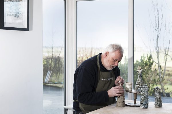 Potter sitting at table painting a piece of pottery. Glass windows and trees are in the background.