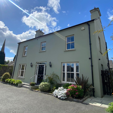5 bedroom house painted a pale green with a black door. There is plenty of greenery around the front of the house and it is pictured on a beautiful sunny day.