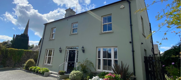 5 bedroom house painted a pale green with a black door. There is plenty of greenery around the front of the house and it is pictured on a beautiful sunny day.