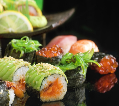 Close-up of assorted sushi rolls topped with avocado, sesame seeds, and fish, served on a black plate with garnish.
