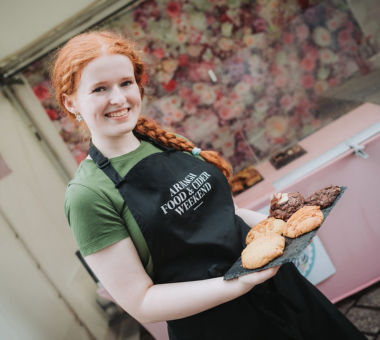 Woman wearing an Armagh Food and Cider Weekend apron holding a tray of baked goods at a market stall.