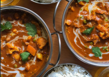 Two bowls of rich curry garnished with herbs, served alongside white rice on a wooden table.