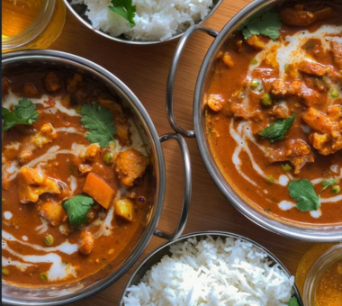 Two bowls of rich curry garnished with herbs, served alongside white rice on a wooden table.