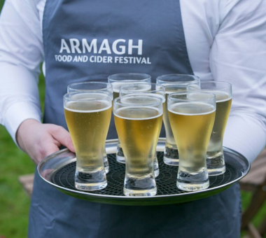 Tray of small cider glasses being served by a person wearing an “Armagh Food and Cider Festival” apron.