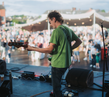 Person playing an electric guitar on stage in front of a large outdoor crowd.