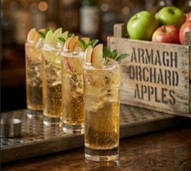 Row of apple cocktails garnished with slices, beside a crate labeled “Armagh Orchard Apples.”