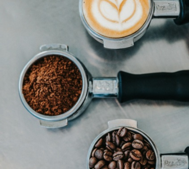 Top-down view of a coffee tasting setup with a portafilter filled with ground coffee, a second portafilter holding whole beans, and a cup of latte with heart-shaped foam art on a metal surface.