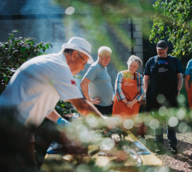 Person demonstrating outdoor cooking over a wooden table while a small group watches nearby.