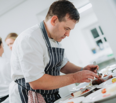 Chef plating a fine dining dish in a bright professional kitchen.