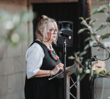 Woman speaking at a microphone while reading from notes at a lectern during an Flash Fiction event.