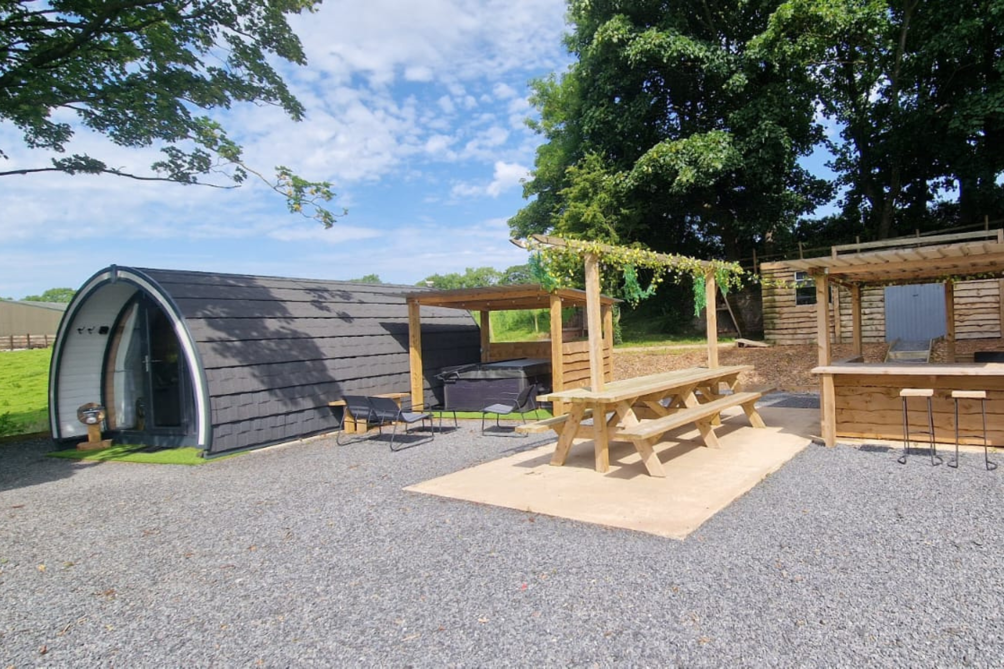 Hawthorn Hideaways Modern glamping pod and outdoor seating area with wooden pergolas and picnic bench on a gravel site.