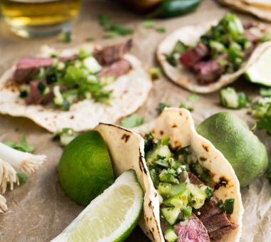 Soft tacos filled with chopped steak, fresh herbs, and vegetables, served with lime wedges on a rustic surface.