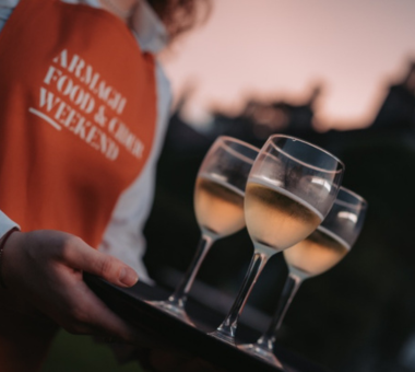 Person wearing an “Armagh Food and Cider Weekend” apron carrying a tray of white wine glasses outdoors