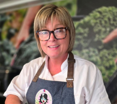 Woman wearing glasses and an apron working in a kitchen setting with food display in the background.
