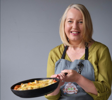 Person holding a frying pan of food while wearing an apron in a studio kitchen setting.