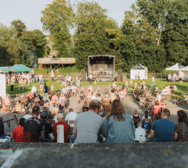 Outdoor crowd facing a stage at a live event with trees in the background.