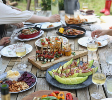 Outdoor table set with tapas-style dishes, including skewered bites, melon with ham, breaded foods, and glasses of white wine.