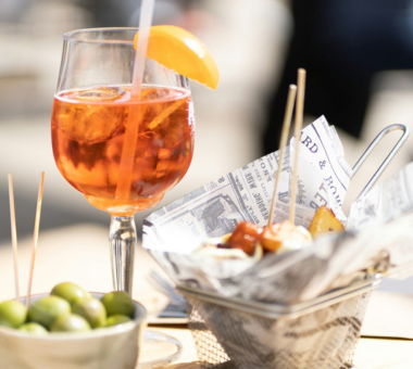 Glass of orange cocktail garnished with a slice, served with tapas and olives on a table.