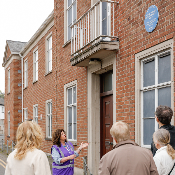 Guide pointing to a blue plaque on a brick building while leading a walking tour group.