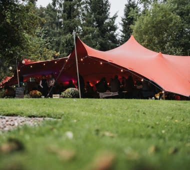 Large stretch tent set up outdoors with people seated inside at a gathering on a grassy lawn.