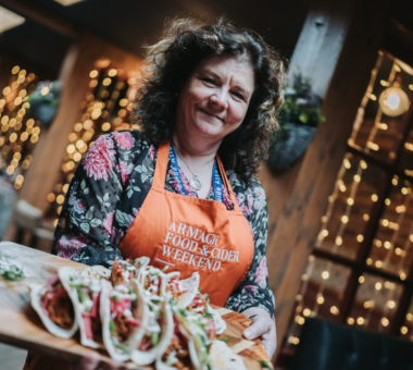 Tour guide Donna Fox wearing an “Armagh Food & Cider Weekend” apron presenting a wooden board of loaded tacos in a warmly lit setting with fairy lights in the background.