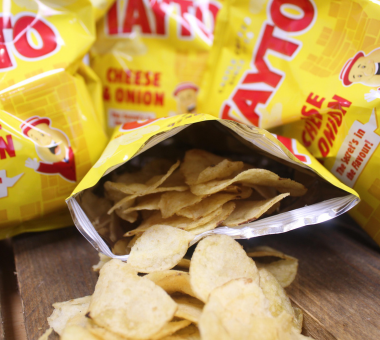 Open bag of Tayto cheese and onion crisps spilling onto a wooden surface, with additional packets in the background.