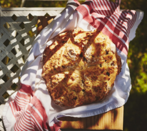 Rustic homemade soda bread wrapped in a striped cloth, resting on a wooden surface outdoors in sunlight.