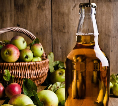 Bottle of cider beside a basket and scattered apples on a rustic wooden surface.