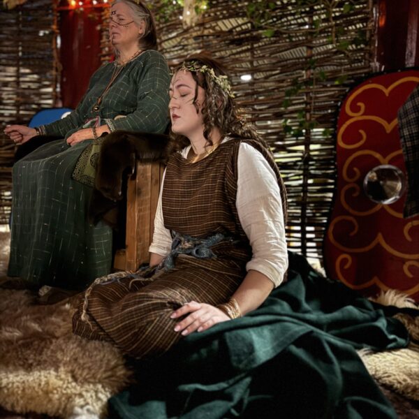 Samhain Two costumed performers seated inside a reconstructed Celtic-style dwelling at Navan Fort, taking part in a historical interpretation surrounded by woven wood walls, animal hides and traditional props.