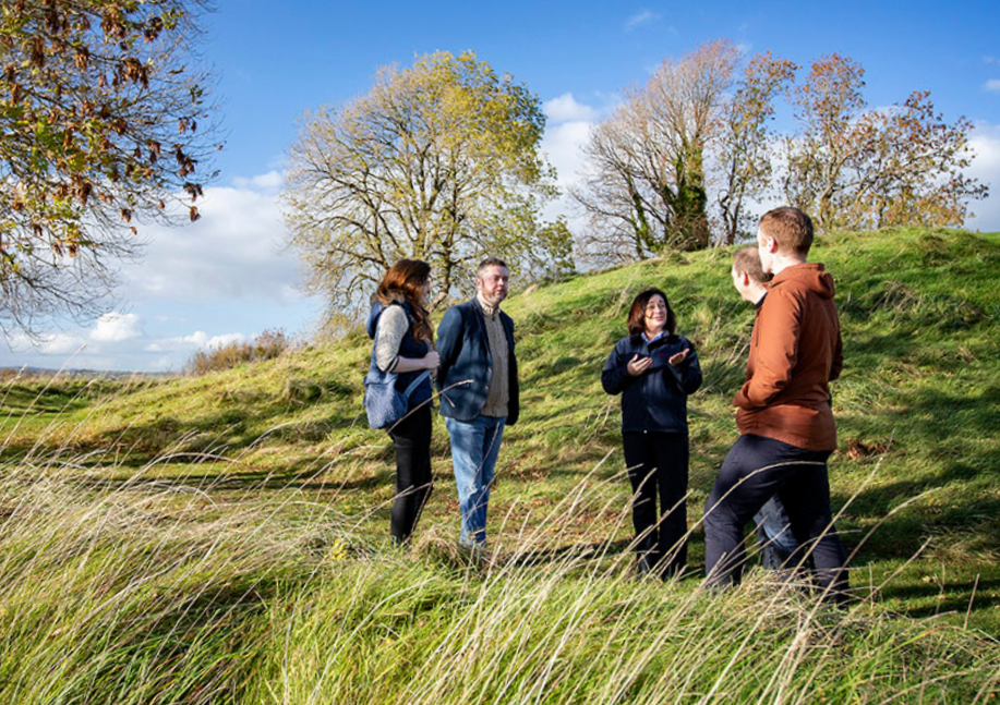 Groups & Travel Trade (Navan Fort) Group taking part in a guided tour below Navan Fort, standing on a grassy slope beside the ancient ceremonial site.